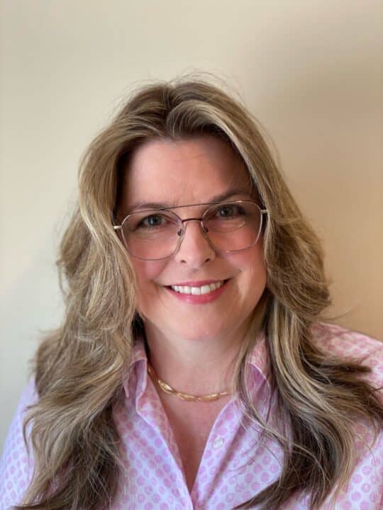 Linda Grgurich, seen from the chest up, wearing a purple blouse, glasses, long blonde hair, and smiling