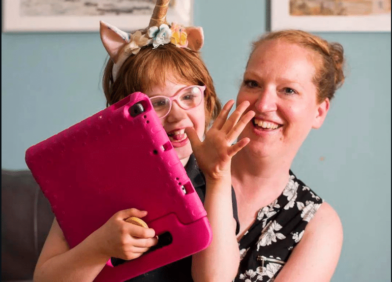 Amelia, a young girl with glasses and red hair, holds her pink ipad and smiles with her mom