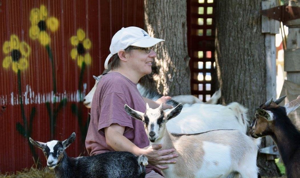 Chey, wearing a purple shirt and white baseball cap, kneels on the ground and is surrounded by goats