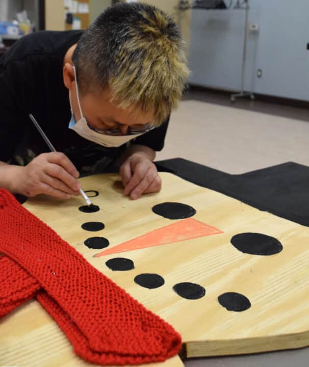 a man in a mask is painting a wooden board. The board has a red scarf around it and is being painted to look like a snowman.