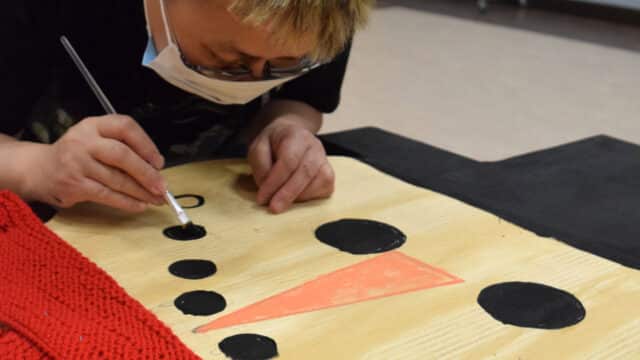 a man in a mask is painting a wooden board. The board has a red scarf around it and is being painted to look like a snowman.