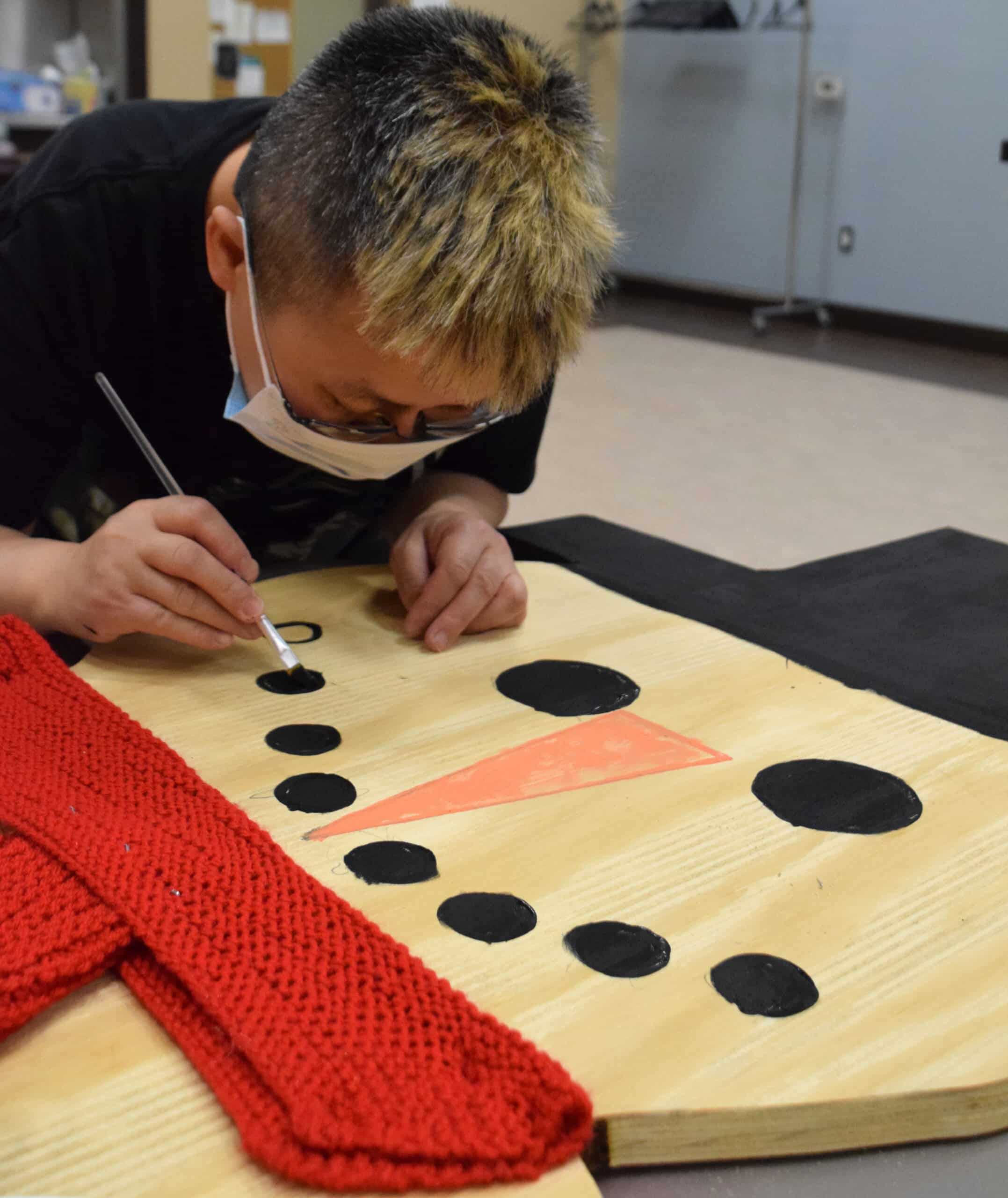 a man in a mask is painting a wooden board. The board has a red scarf around it and is being painted to look like a snowman.