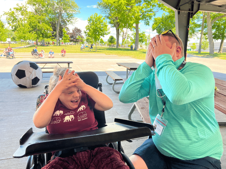 Tyler (a young man in a wheelchair) and Courtney (a young woman) sit together and cover their faces in laughter