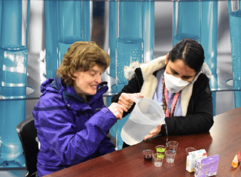 Two people are sitting at a table doing a science experiment. In the back is an edited photo of test tubes filled with blue liquid
