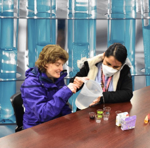 Two people are sitting at a table doing a science experiment. In the back is an edited photo of test tubes filled with blue liquid
