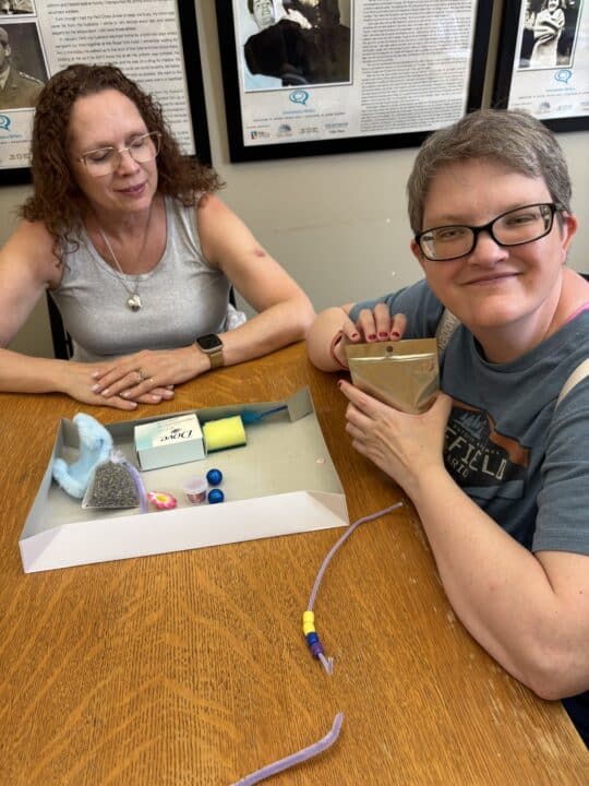 Two women are sitting at a table. There is a box in front of them with different tactile and sensory friendly materials.