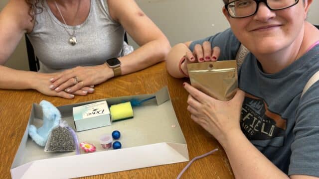 Two women are sitting at a table. There is a box in front of them with different tactile and sensory friendly materials.