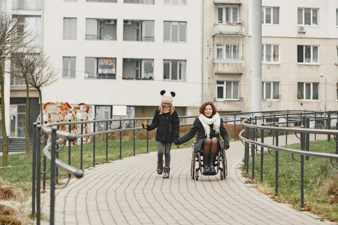 A women in a wheelchair is holding hands with a women. The two women are outside on a paved path. In the background appears to be post secondary residence/ a school.