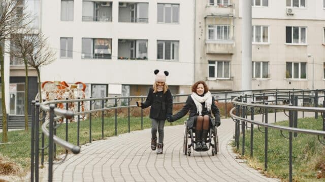 A women in a wheelchair is holding hands with a women. The two women are outside on a paved path. In the background appears to be post secondary residence/ a school.