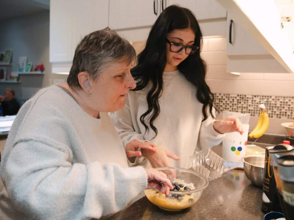 An older woman and a young woman in a kitchen, working together to measure ingredients into a silver mixing bowl.
