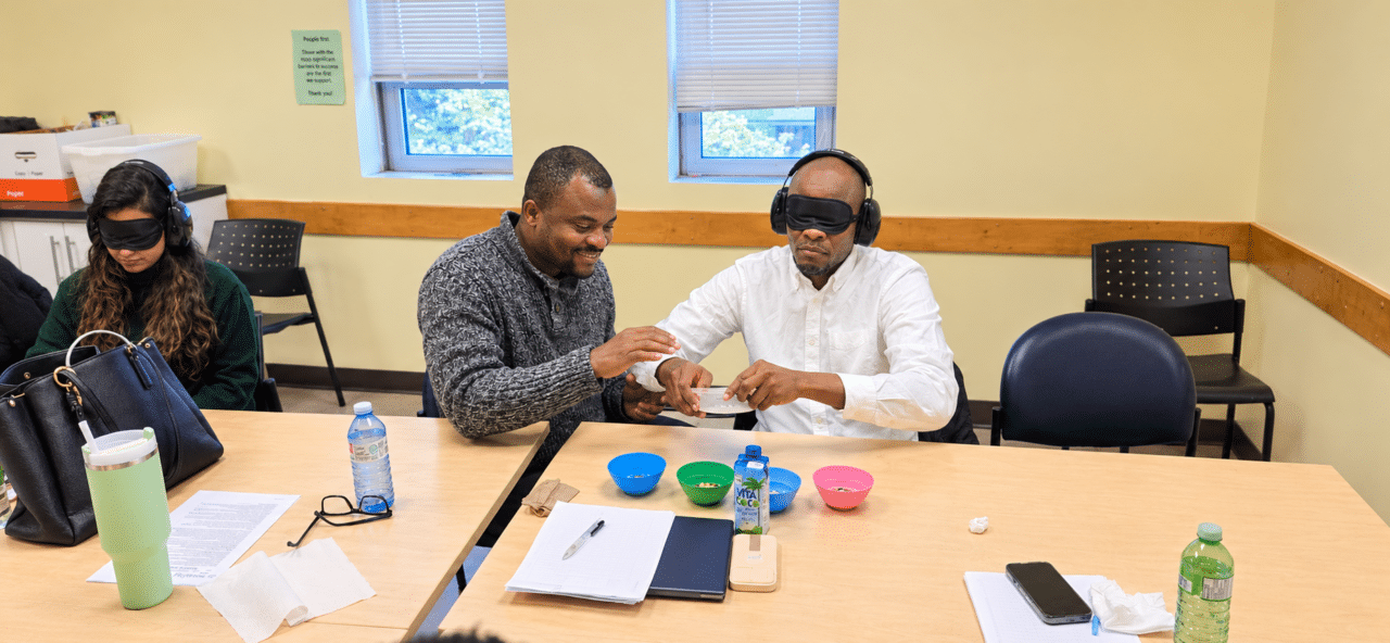Two participants take part in a sensory awareness exercise at a table, with one wearing a blindfold and headphones while another guides his hands as they handle materials. A second participant wearing a blindfold and headphones sits nearby. Bowls, papers and other items are arranged across the table in a community training room.
