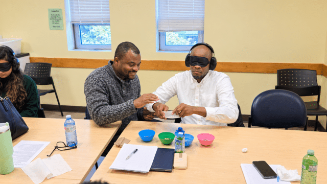 Two participants take part in a sensory awareness exercise at a table, with one wearing a blindfold and headphones while another guides his hands as they handle materials. A second participant wearing a blindfold and headphones sits nearby. Bowls, papers and other items are arranged across the table in a community training room.