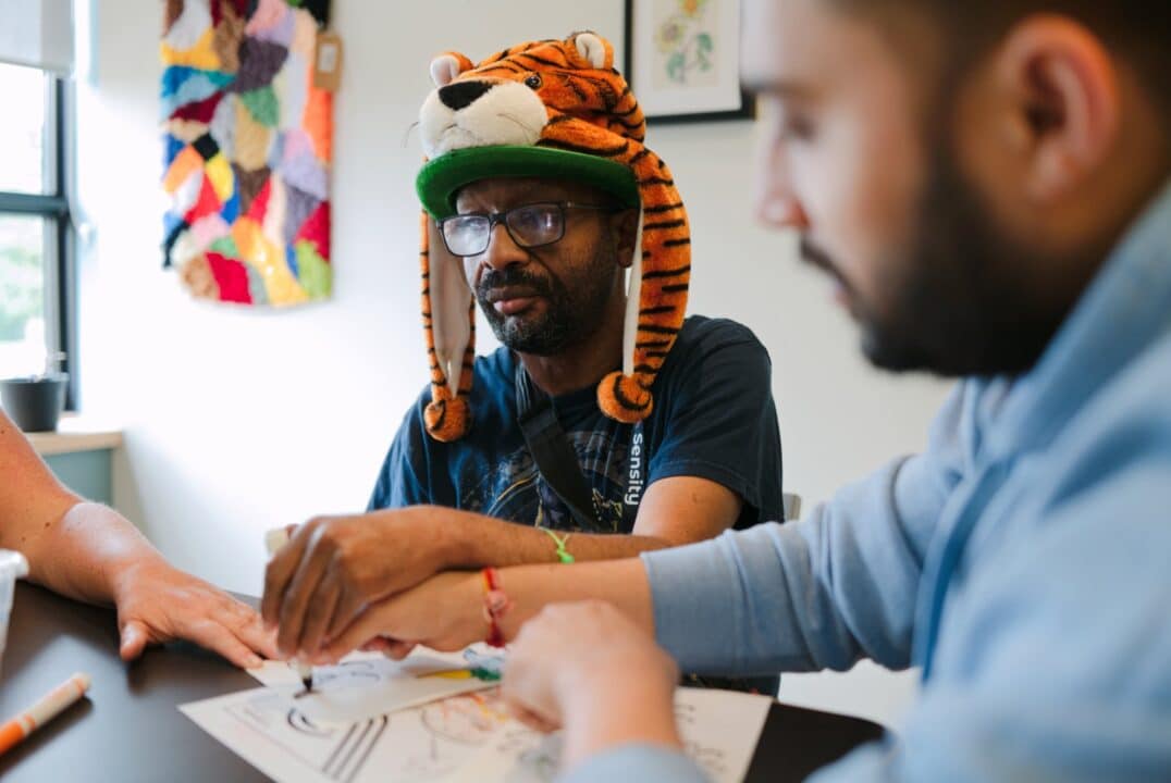 A man wearing a hat sitting at a table and drawing on paper with the guidance of an intervenor.